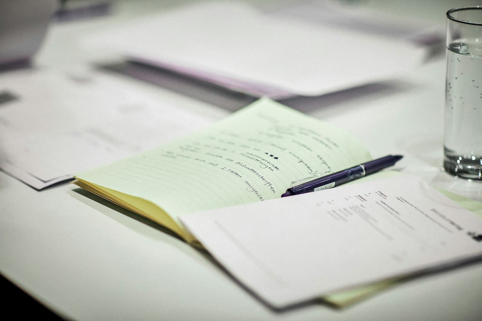 A legal pad covered in handwritten notes with a pen laid across it, beside printed pages and a glass of water.