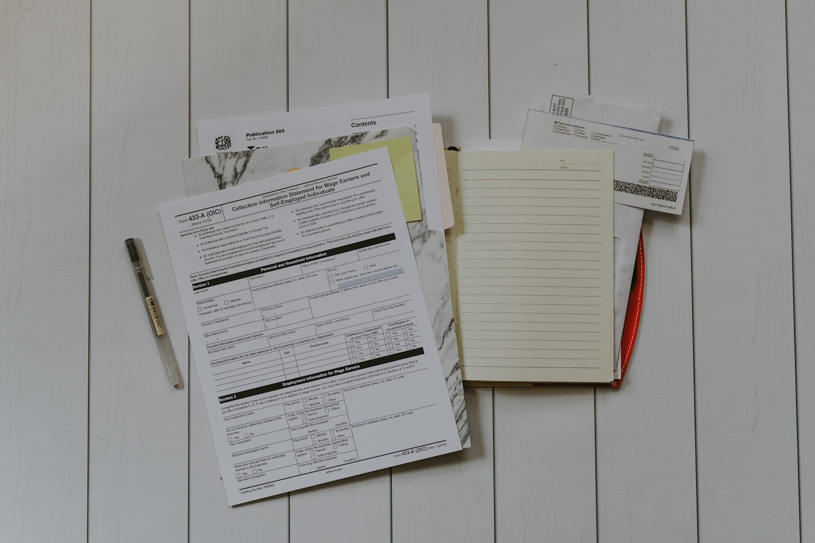 Printed forms and statements spread across a white table beside an open notebook and a pen.