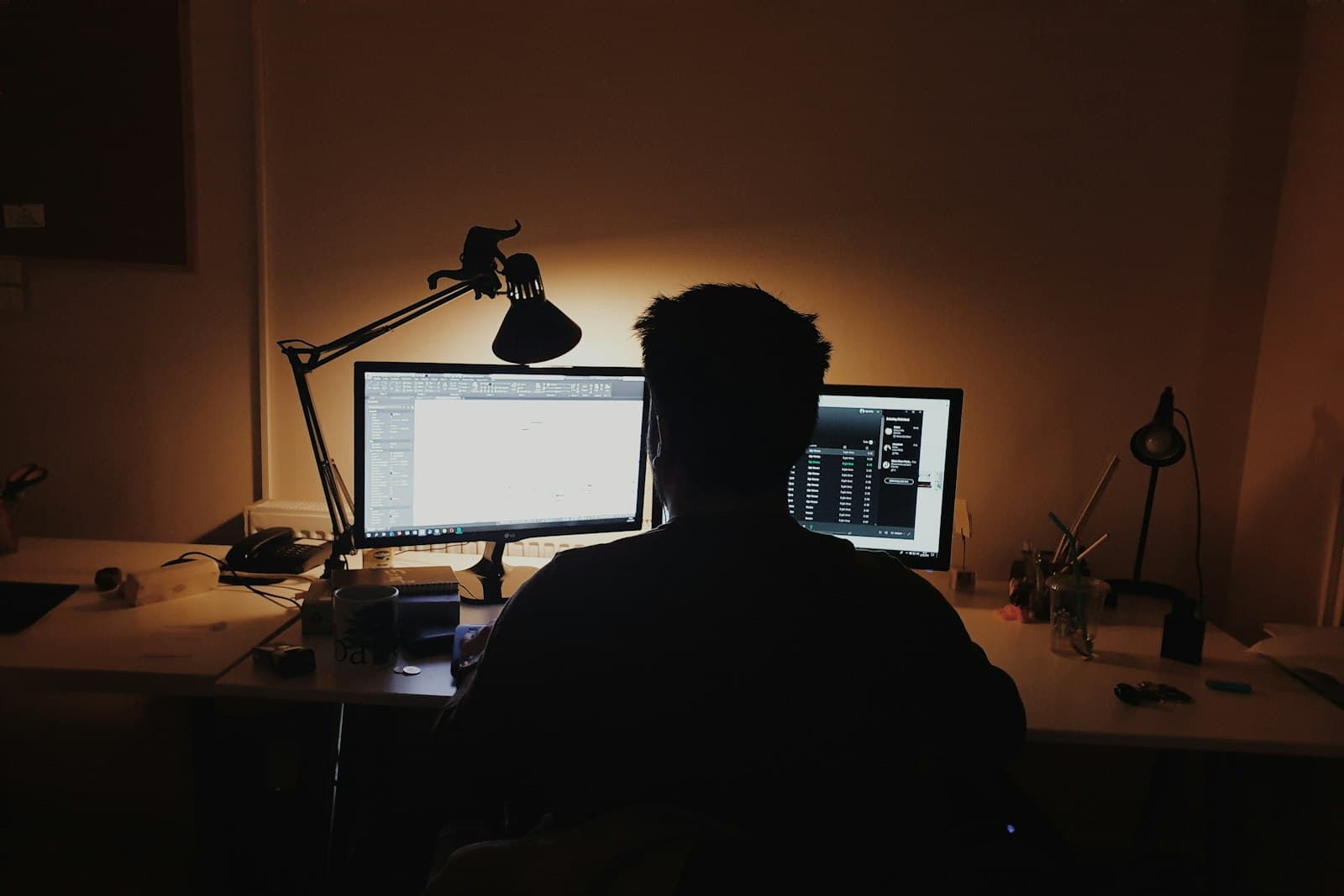 A person silhouetted at a desk in a dark room, lit by two monitors and a warm desk lamp.