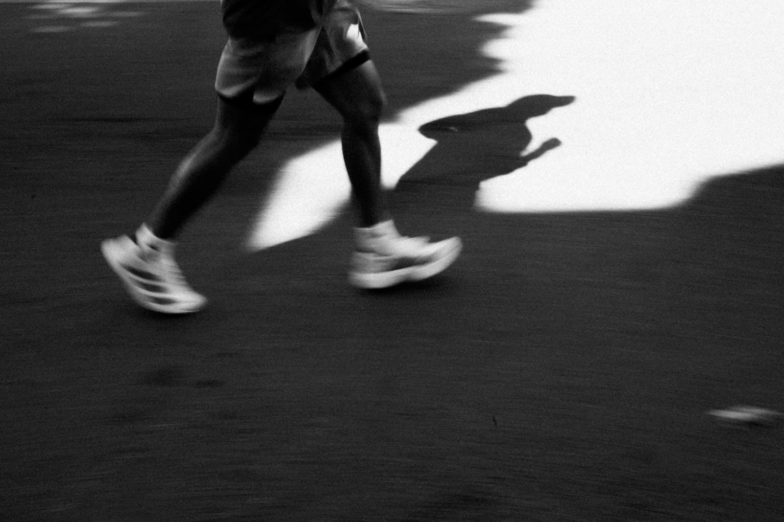 A black and white photograph of a runner's legs mid-stride on asphalt, casting a long shadow across the road.
